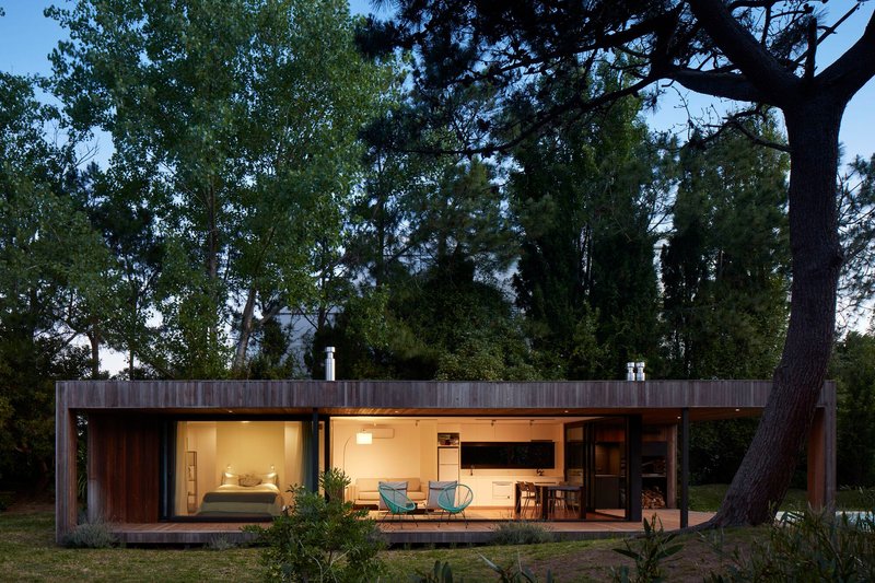 Long timber volume with continuous glazing and lit interior at dusk in a wooded clearing