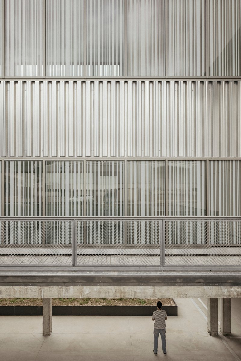 Vertical metal screen facade with mesh balustrade viewed from concrete-columned ground-level courtyard with visitor