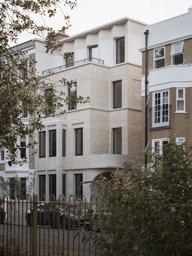 Street view of the pale brick facade with vertical windows nestled between adjacent buildings and greenery