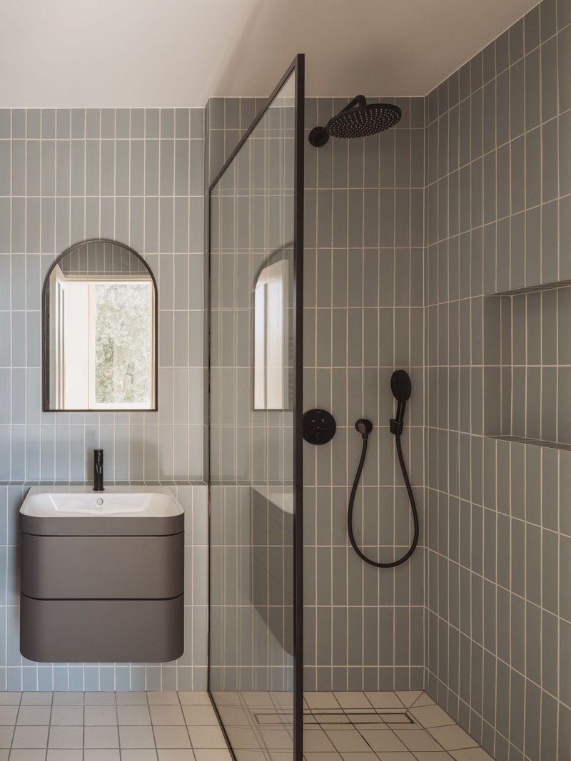 Bathroom interior with olive vertical tile shower enclosure, arched mirror and wall-mounted vanity under soft daylight