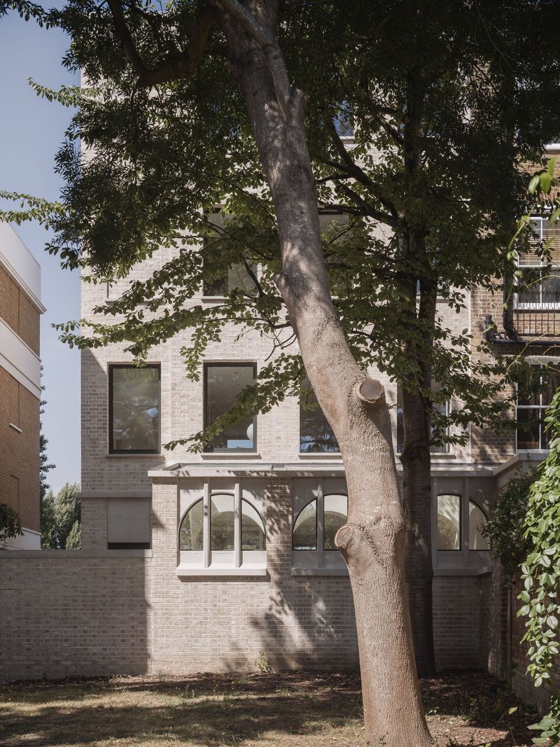 Brick facade with arched ground-floor openings and square windows framed by a large tree casting dappled shadows