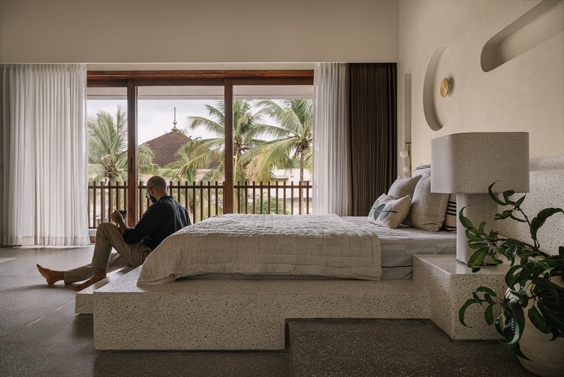 Bedroom with person seated on terrazzo bed platform overlooking palm trees through glazed opening