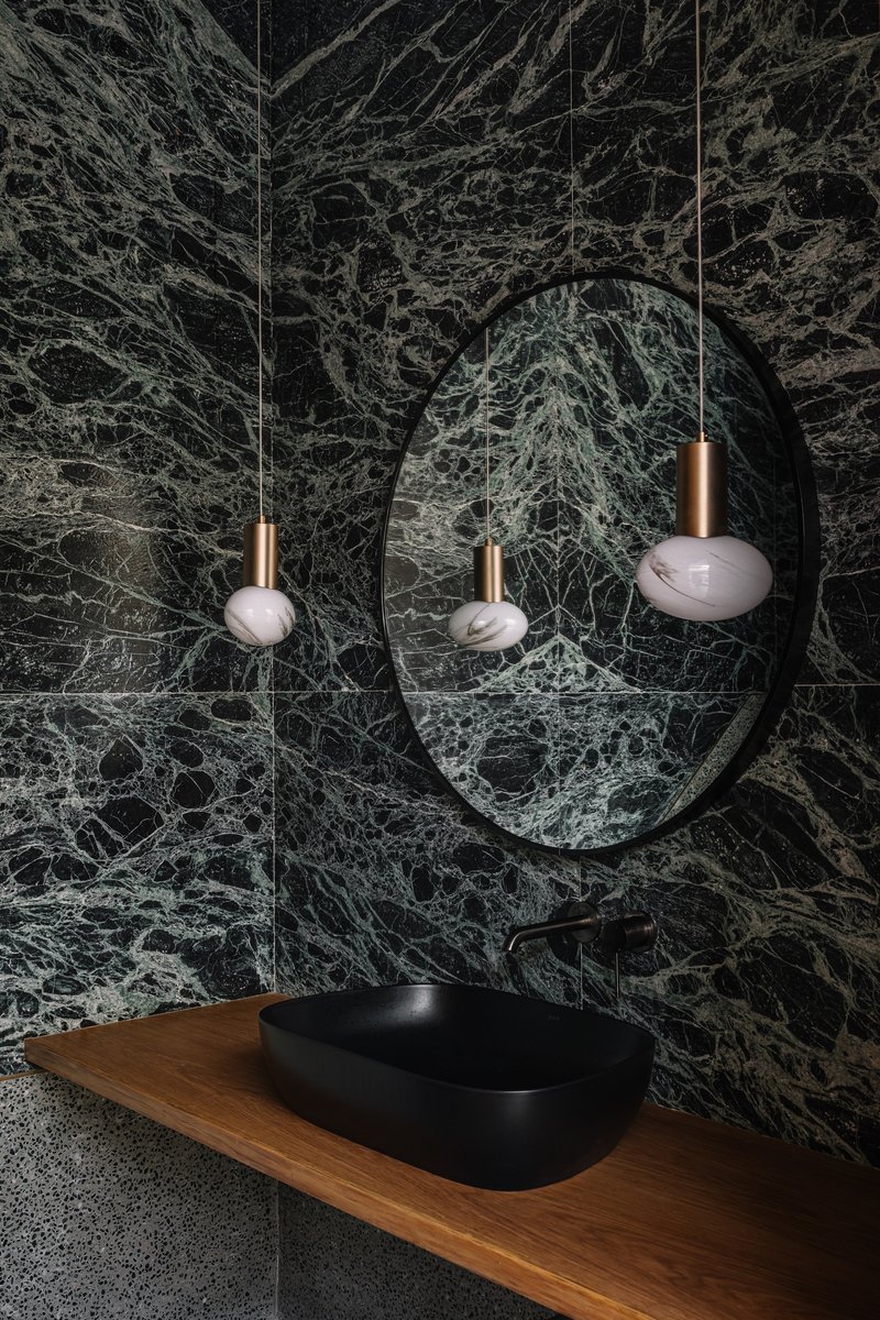 Round mirror reflecting pendant lights above a timber vanity with black basin against green marble