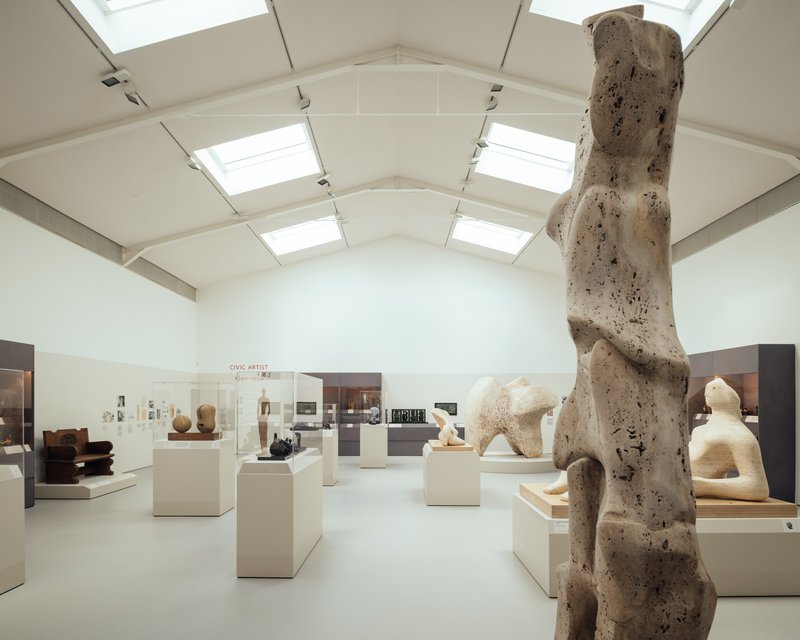 Gallery space with vaulted ceiling, skylights, and sculpture displays on white plinths