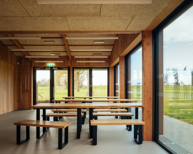 Communal dining area with timber benches and tables overlooking a grassy field through glazed doors