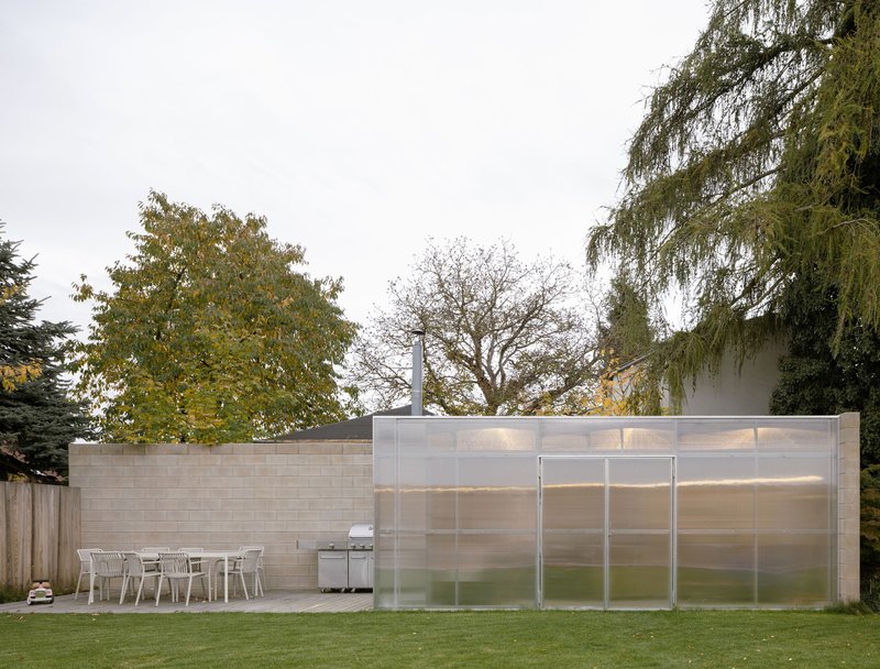 Outdoor dining terrace with concrete paving beside translucent polycarbonate wall and weathered timber fence under overcast sky
