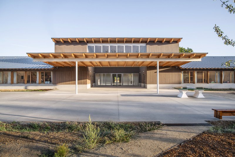 Main entrance with layered timber canopies and white columns sheltering a concrete plaza at dusk