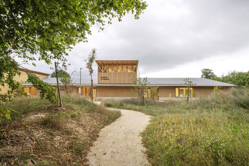Gravel pathway through wildflower meadow leading to low timber and earthen volumes under overcast sky