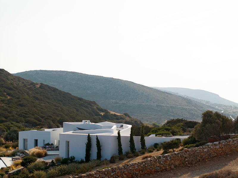 Side view of the white volumes with cypress trees against the mountain backdrop at dusk