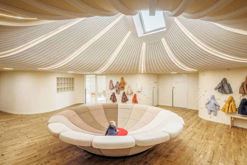 Interior playroom with upholstered circular seating platform beneath a sculptural timber ceiling and central skylight