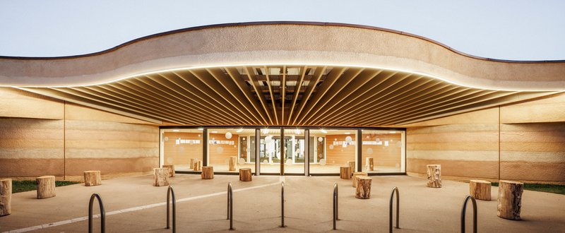 Evening view of the illuminated entrance with exposed timber rafters beneath the curved rammed earth canopy