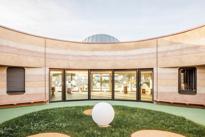 Circular courtyard with curved rammed earth walls, glazed doors and a white sphere on artificial turf