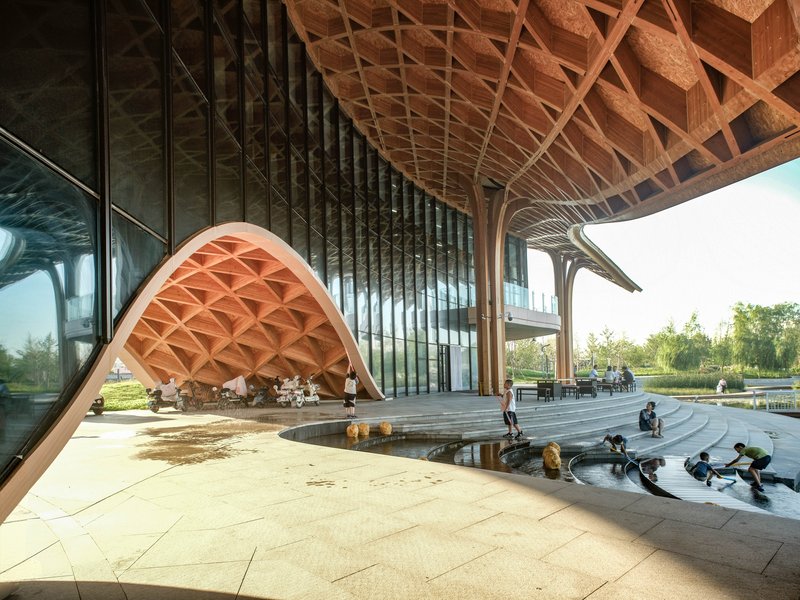 Sheltered plaza with water feature where children gather beneath the coffered timber roof and reflective glass curtain wall