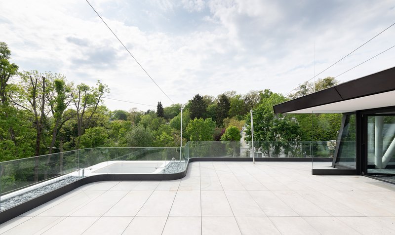 Rooftop terrace with pale pavers, glass balustrade, and tree canopy under cloudy sky
