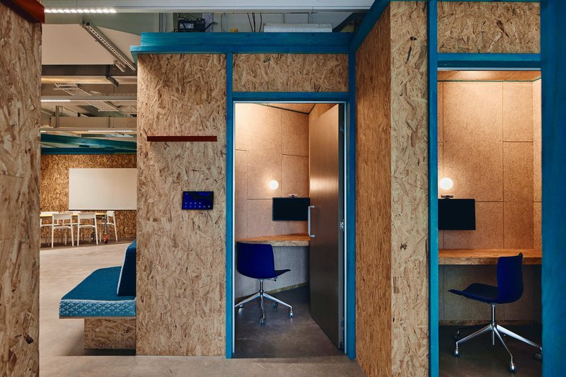 Individual work booths framed in blue steel with oriented strand board and cork paneling under exposed ceiling