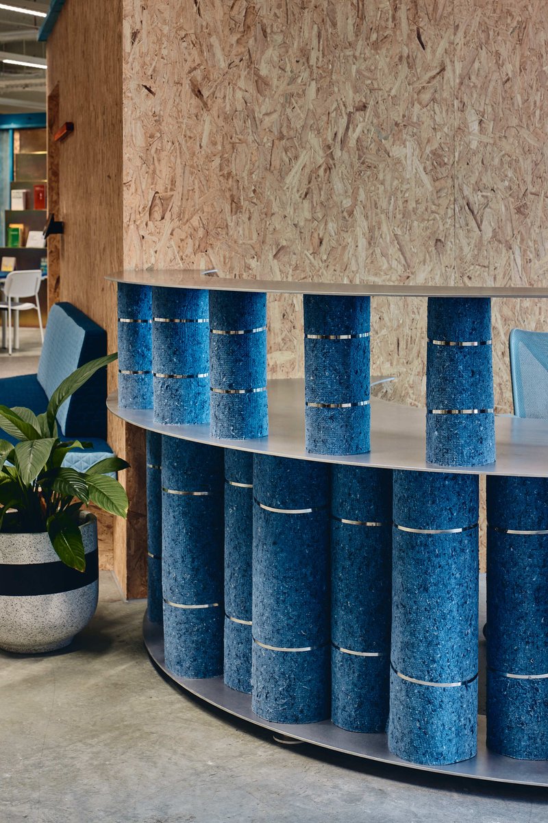 Curved reception desk clad with rolled blue felt cylinders in front of oriented strand board wall