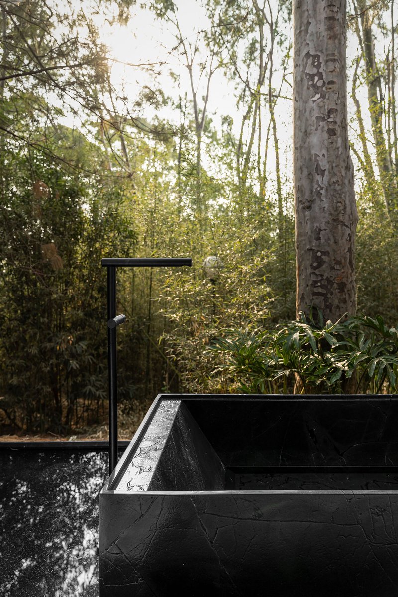 Black concrete outdoor bathtub with wall-mounted faucet surrounded by eucalyptus trees in dappled afternoon light