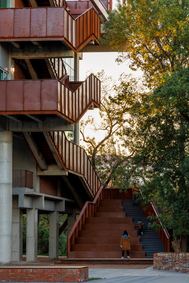 Wide exterior staircase with weathering steel cladding and concrete columns under trees in warm afternoon light