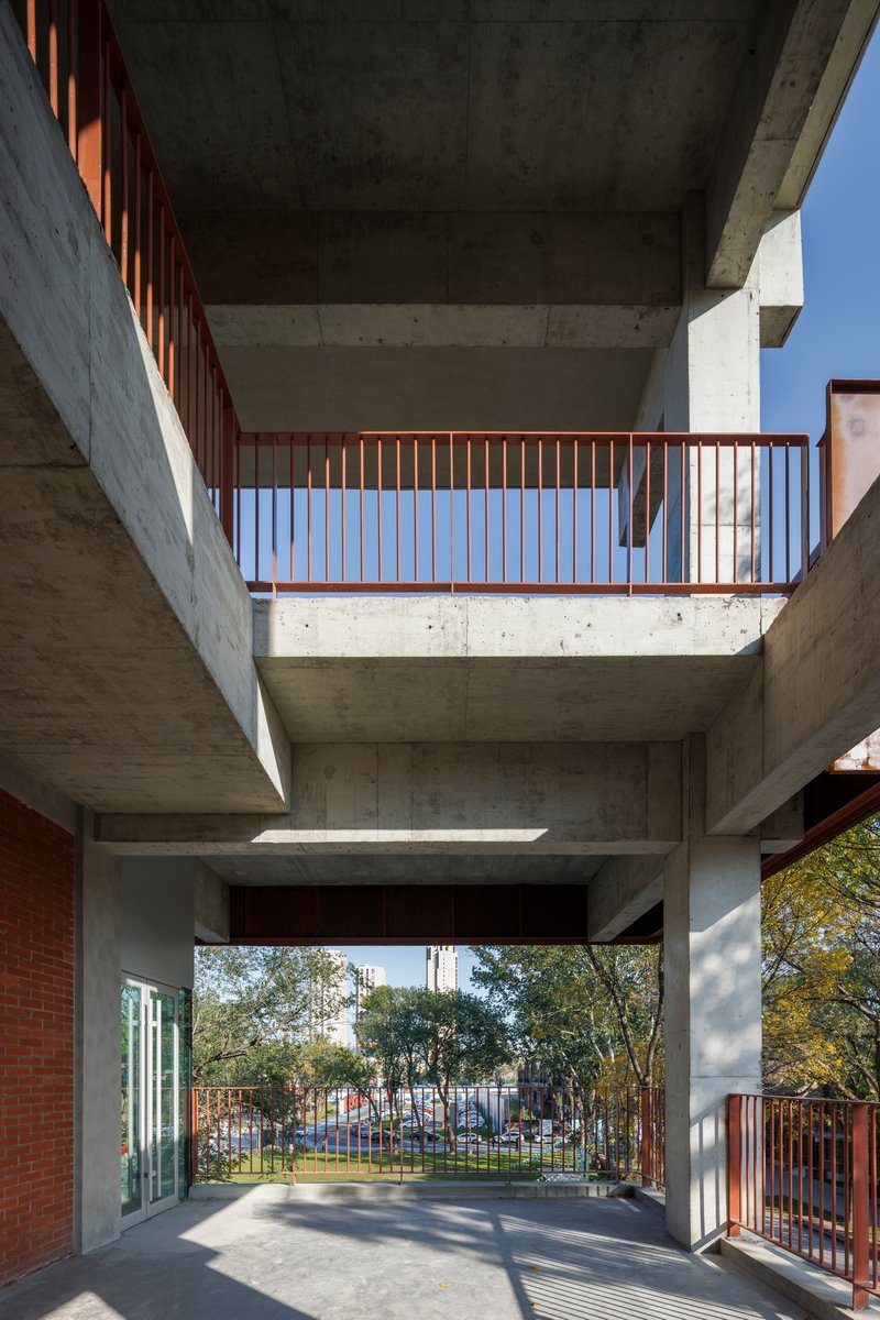 Ground level view through a concrete pilotis passage with steel balcony railings framing a planted courtyard beyond
