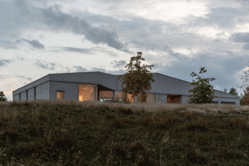 Metal-clad volumes with illuminated glazing at dusk viewed from a sloping meadow with trees