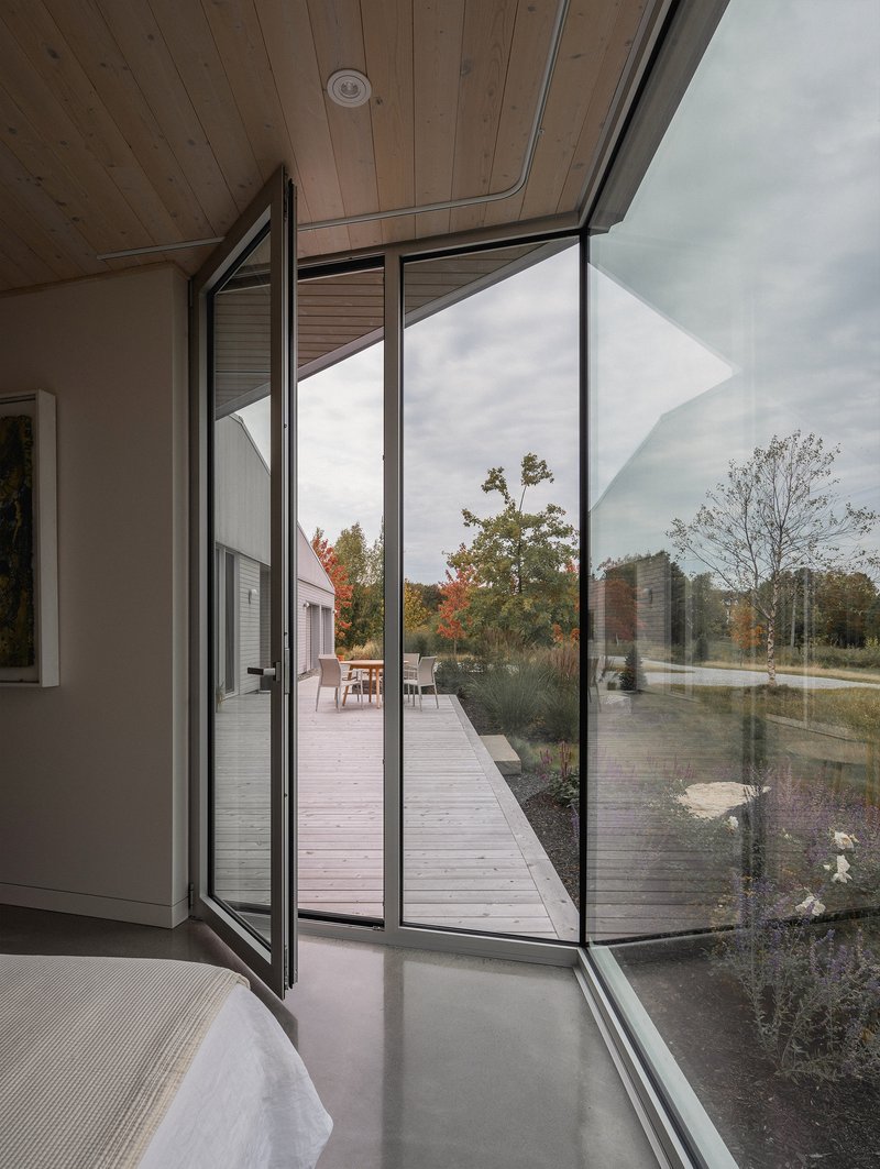 Pivoting glass door opens from bedroom onto timber deck framed by autumn foliage and cloudy sky