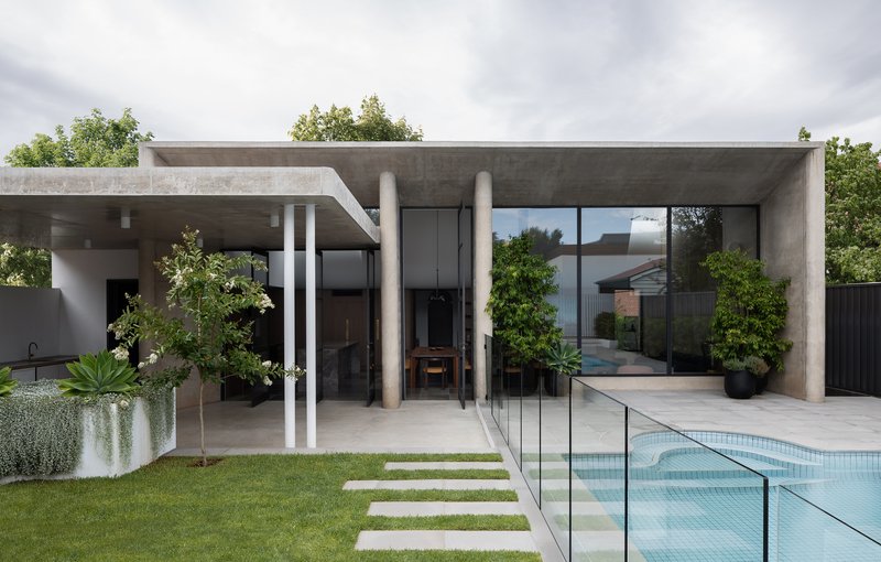 Concrete pavilion with cylindrical columns and full-height glazing overlooking a pool and lawn with stepping stones