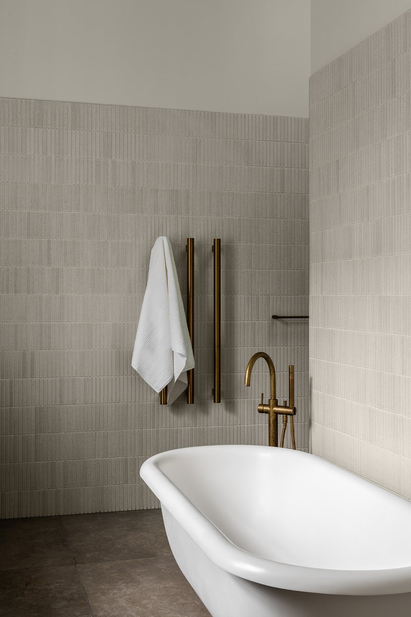 Freestanding white bathtub with brass fixtures against ribbed ceramic tile walls in muted tones