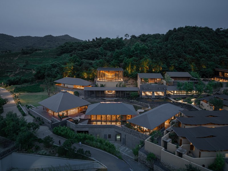 Aerial view of clustered structures with dark pitched roofs nestled into forested hillside at dusk