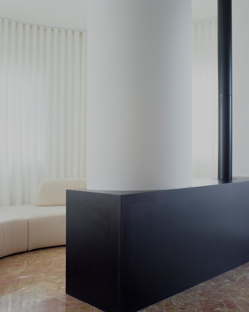 Black steel fireplace hearth next to curved sofa with white curtain and stone floor