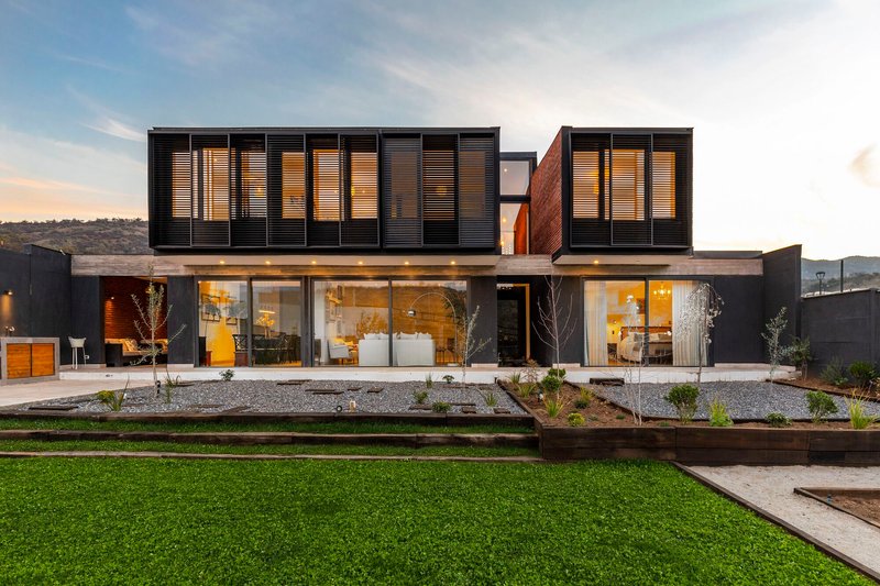 Garden facade with planted beds and floor-to-ceiling glazing beneath slatted black upper volume at twilight