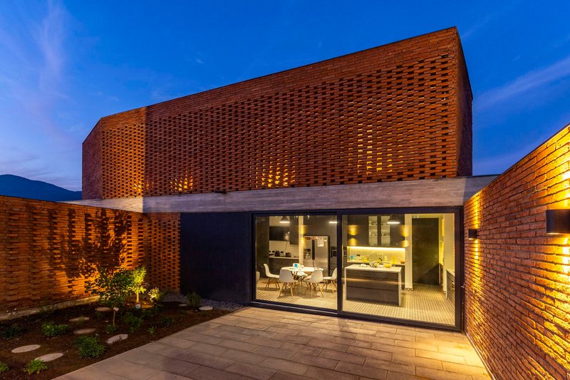 Courtyard view of glazed dining room beneath curved perforated brick volume with planted bed at twilight