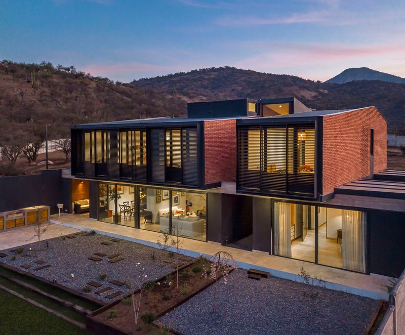 Two-story residence with red brick upper volume and louvered metal facade illuminated at twilight with hillside behind