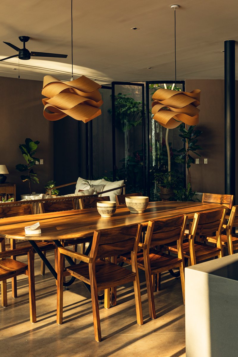 Timber dining table beneath sculptural pendant lights with afternoon sun casting shadows across the concrete floor
