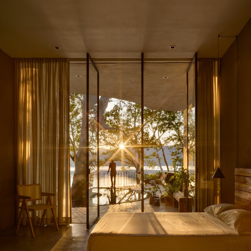 Bedroom with steel-framed glass doors opening to a terrace with a large tree at sunset