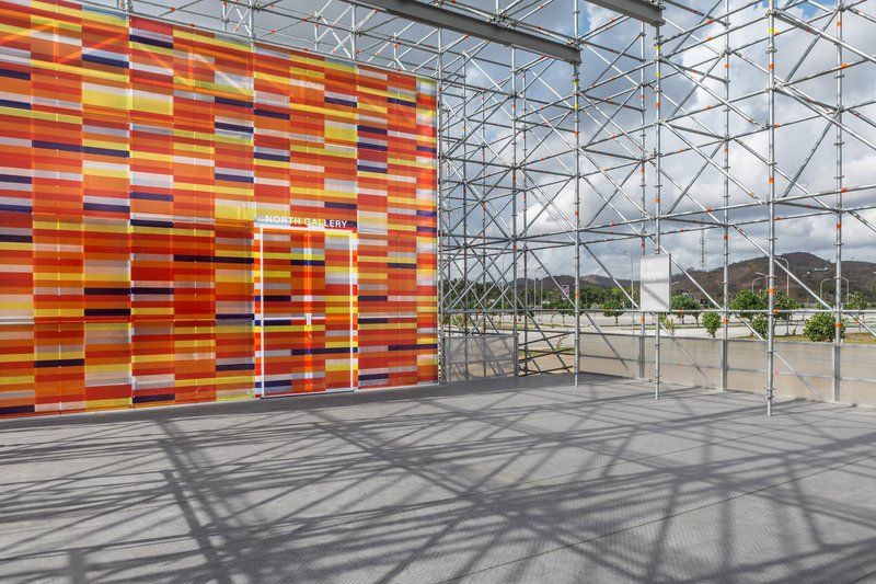 Entry doorway cut through the multicolored horizontal panel wall with scaffolding casting shadows on concrete