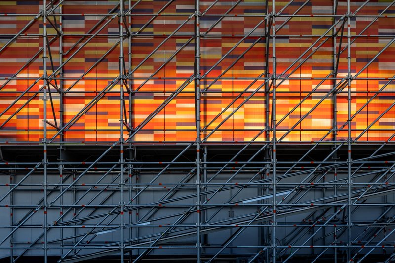 Horizontal bands of orange and warm-toned panels backlit behind the diagonal steel cross-bracing
