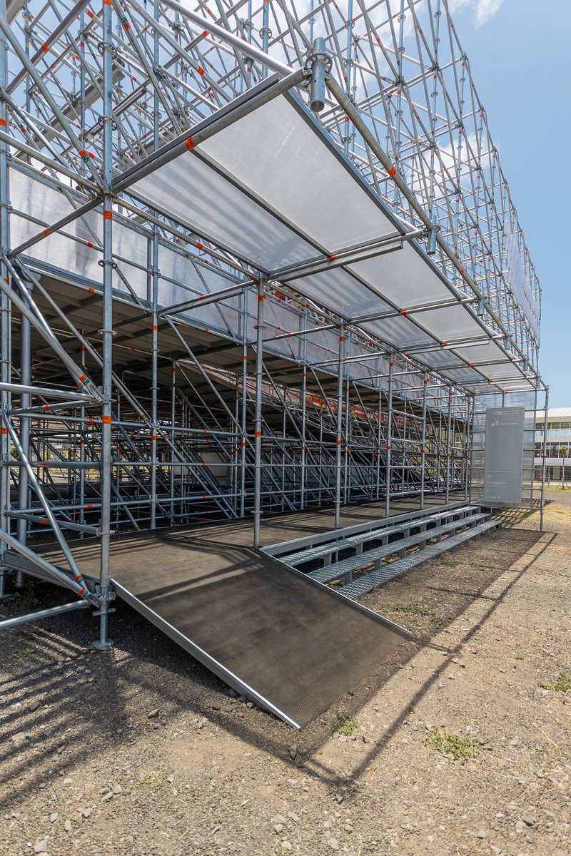 Metal scaffold structure with tensile fabric canopy and accessible ramp under clear blue sky