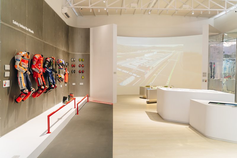 Exhibition gallery with ribbed timber walls displaying racing suits and projection screen under exposed truss ceiling