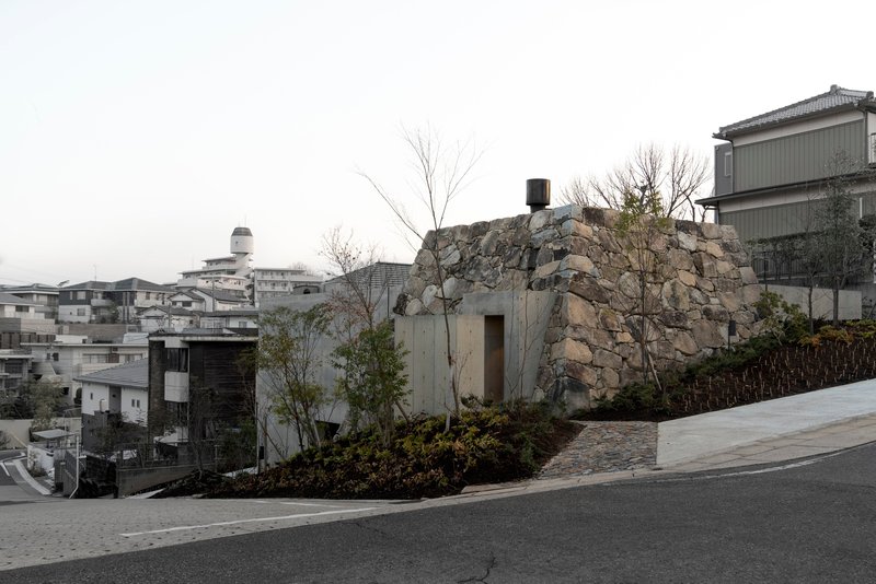 Stone-clad volume nestled into a planted slope along a residential street on an overcast day