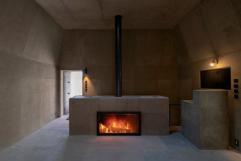 Concrete fireplace block with black flue pipe and glowing fire inside a vaulted room at dusk