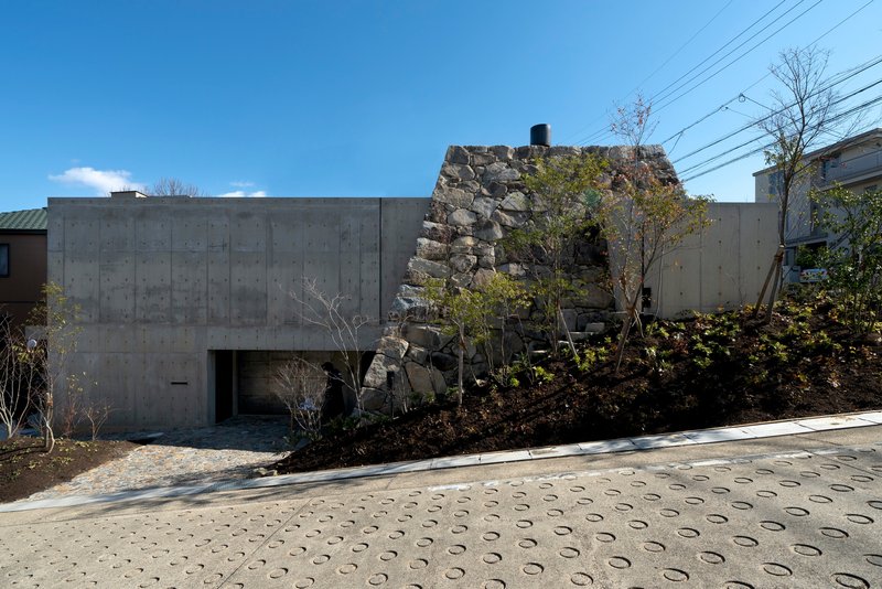 Concrete facade with recessed entrance beside a reconstructed stone wall and paved plaza under blue sky