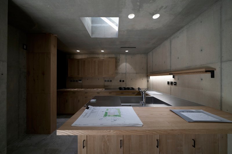 Kitchen with timber cabinetry and board-formed concrete walls illuminated by a central skylight