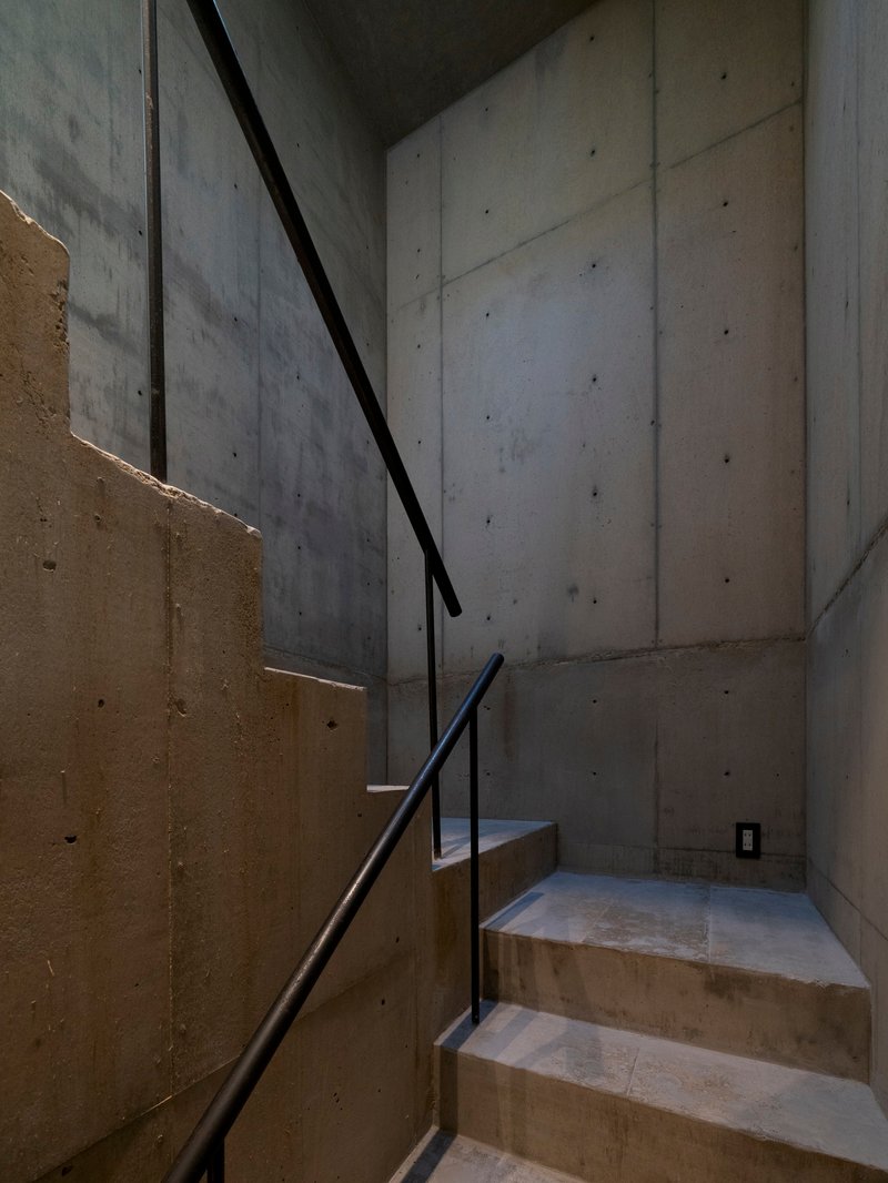 Board-formed concrete staircase with black steel handrail ascending between cast concrete walls