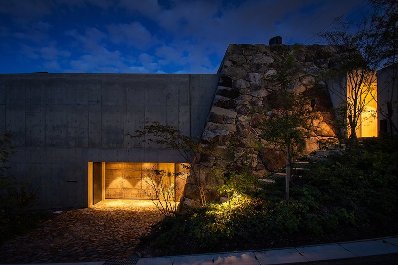 Exterior view at dusk showing glazed entry between concrete wall and stone outcrop with warm interior lighting