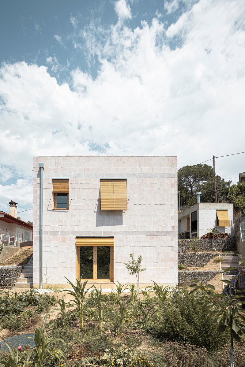 Pale masonry facade with timber-framed windows set among young plantings under a cloudy sky