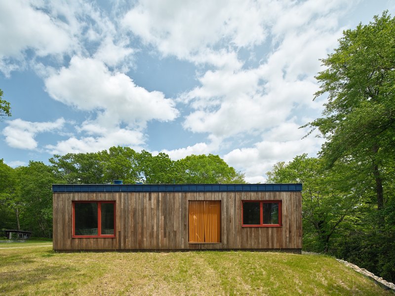 Garden facade showing weathered timber cladding with red-framed windows and vertical slat entry door under blue metal parapet