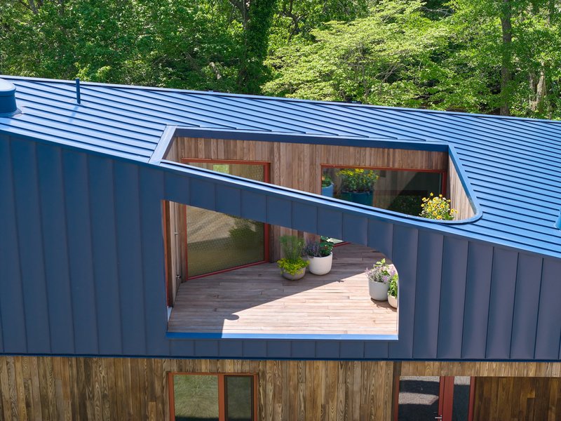 Cutout terrace in blue metal-clad upper volume with timber decking and potted plants amid dense foliage