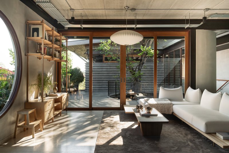 Living room with exposed concrete ceiling, timber-framed glazed doors opening to an interior courtyard with dappled sunlight