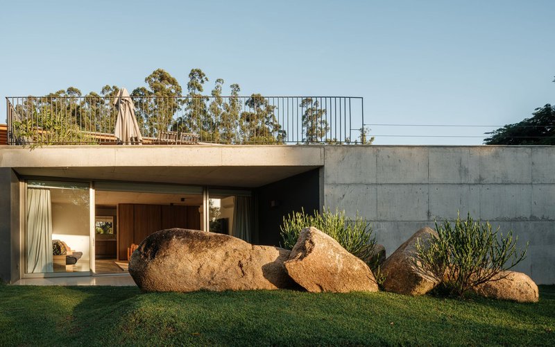 Concrete volume with rooftop garden and large boulders flanking the glass-walled entry in afternoon light