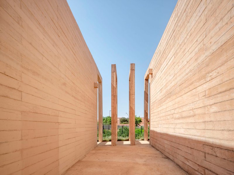 Narrow outdoor passage between rammed earth walls with four free-standing timber columns framing sky